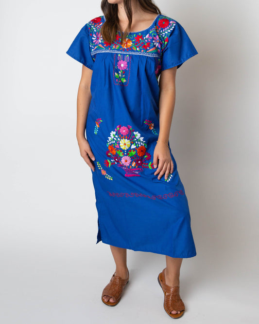 A woman wearing a traditional Mexican blue embroidered dress with colorful floral patterns.