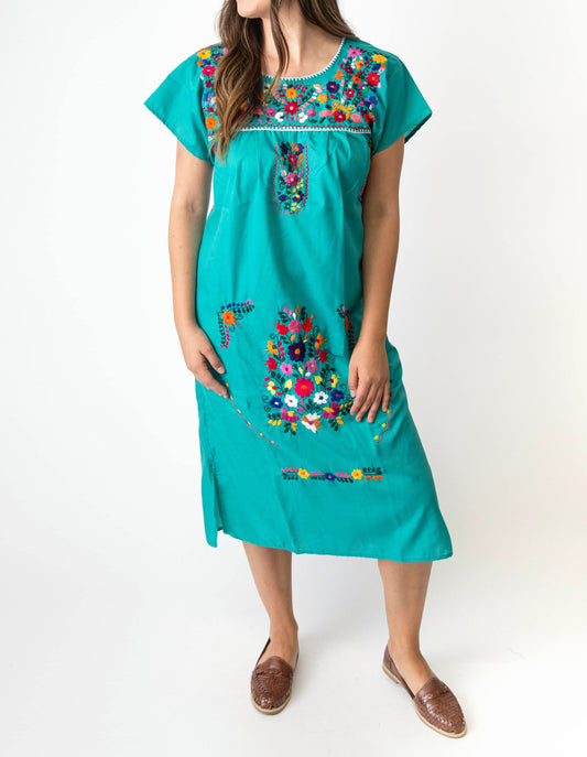 A woman wearing a traditional Mexican embroidered dress in teal color, featuring colorful floral patterns.